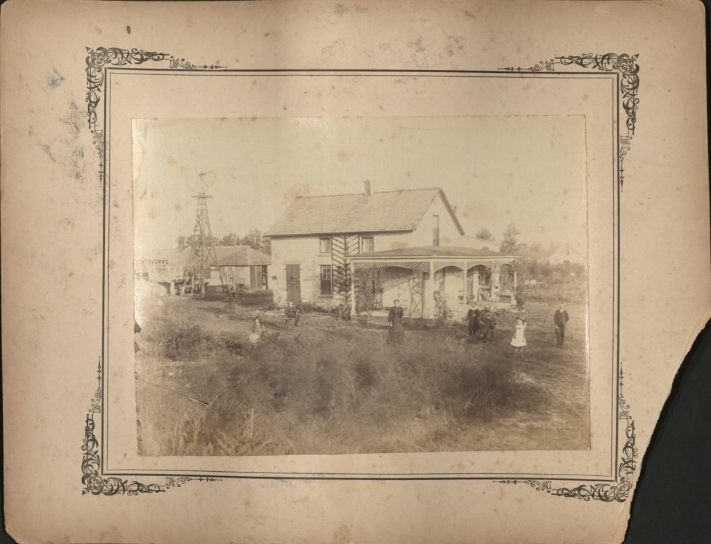 Sepia-toned photograph of family members spaced out in front of a light-colored two-story frame house with a large front porch. Outbuildings and a windmill can be seen in the background.