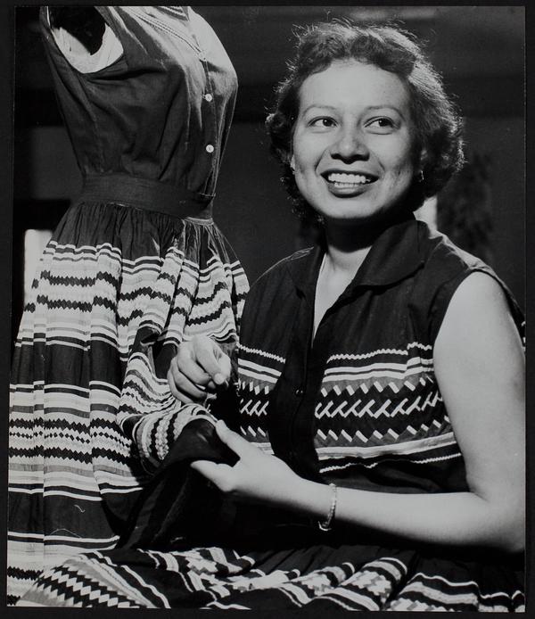 Black-and-white photograph of a smiling woman next to a dress she is working on.