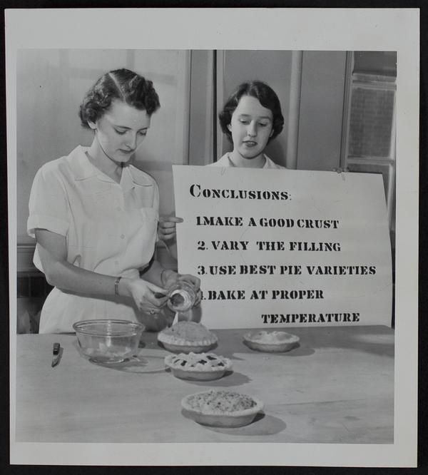 Black-and-white photograph of two women standing behind pies on a table.