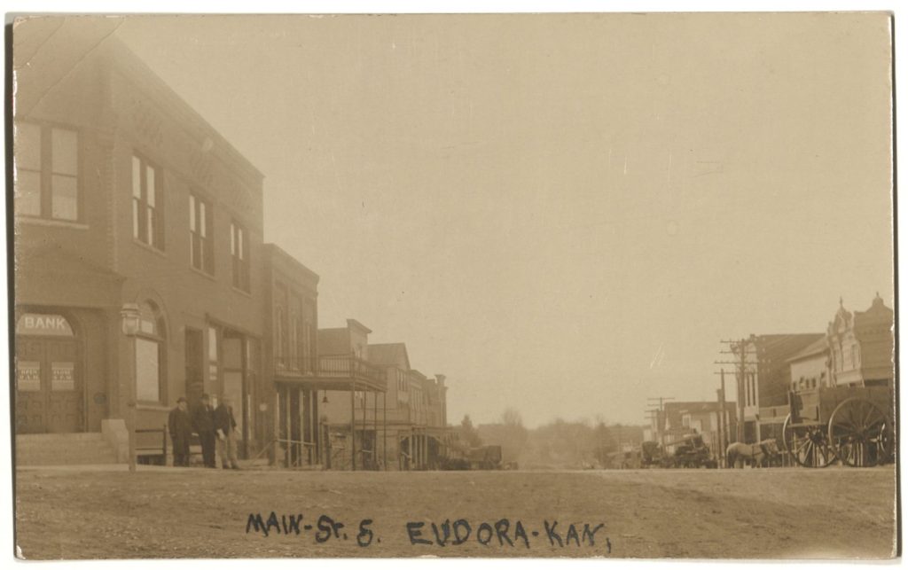 Sepia-toned photo of a dirt street with two-story buildings - and some horses and wagons - on each side. A handwritten note says "Main St S, Eudora, Kan."