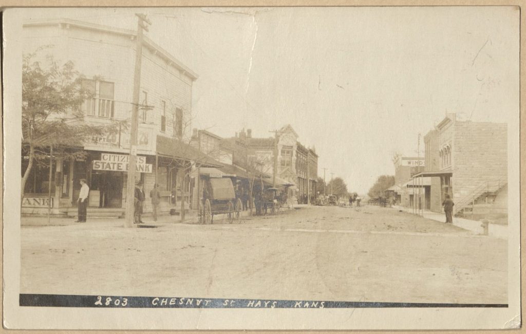 Sepia-toned photo of a dirt street with two-story buildings - and some horses and wagons - on each side. A handwritten note says "2803 Chesnut [sic] St, Hays, Kans."
