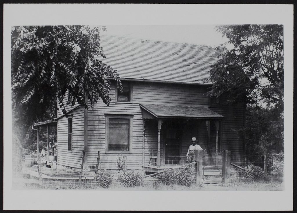 Black-and-white photograph of a man standing in front of a two-story wood frame house with a front porch.
