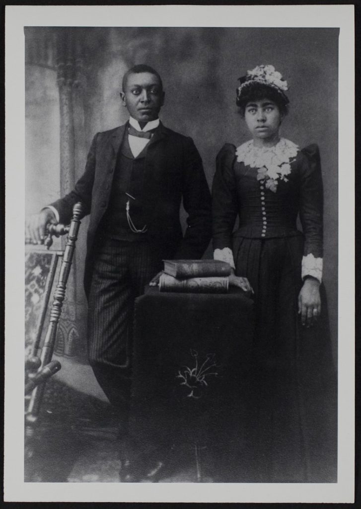 Black-and-white photograph of an African American couple standing together next to a chair and behind a table with books.