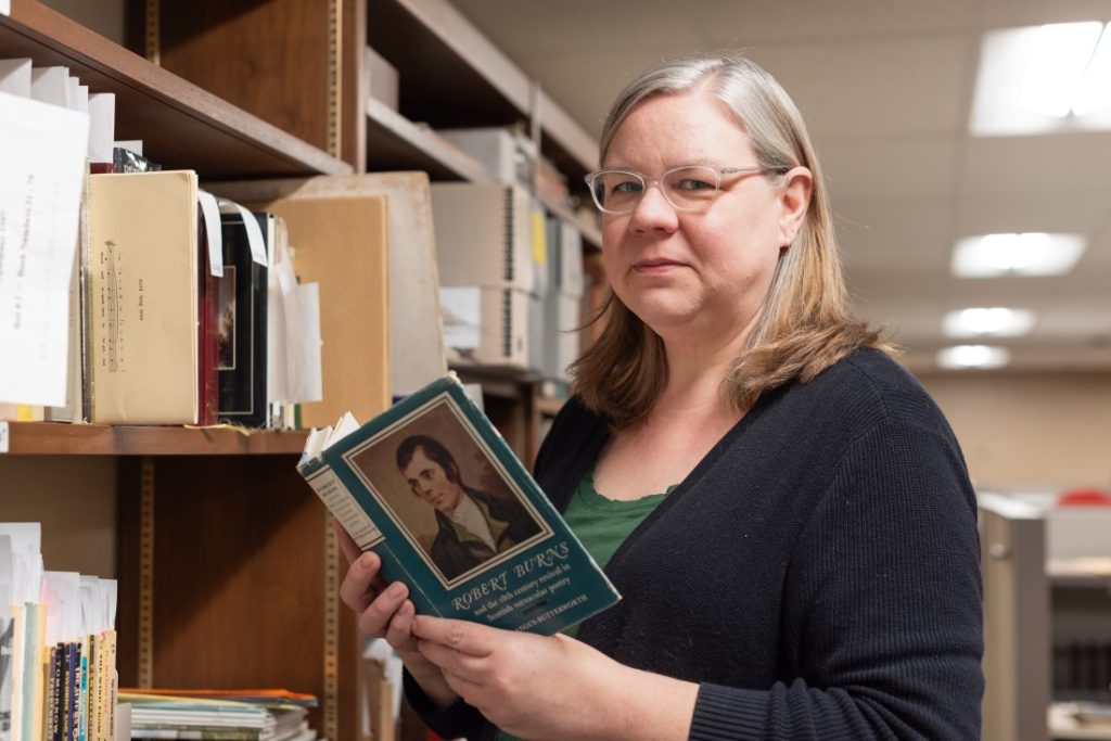Photograph of a woman standing in front of a wooden bookshelf. She is holding a book about Robert Burns.