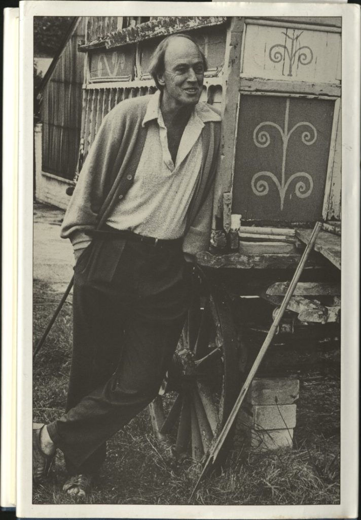 Black-and-white photograph of an adult Roald Dahl standing and leaning against a decorated wagon.
