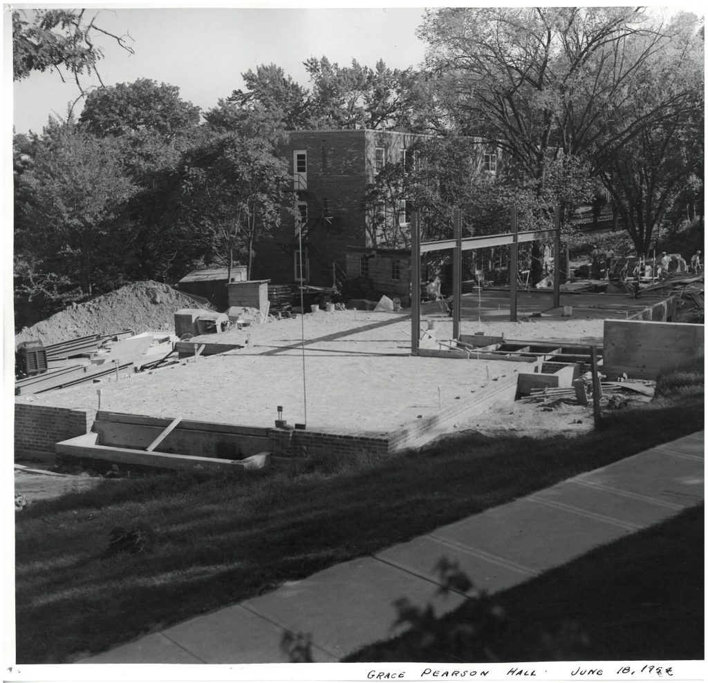 Construction of a concrete foundation for a building, with a brick building in the background.