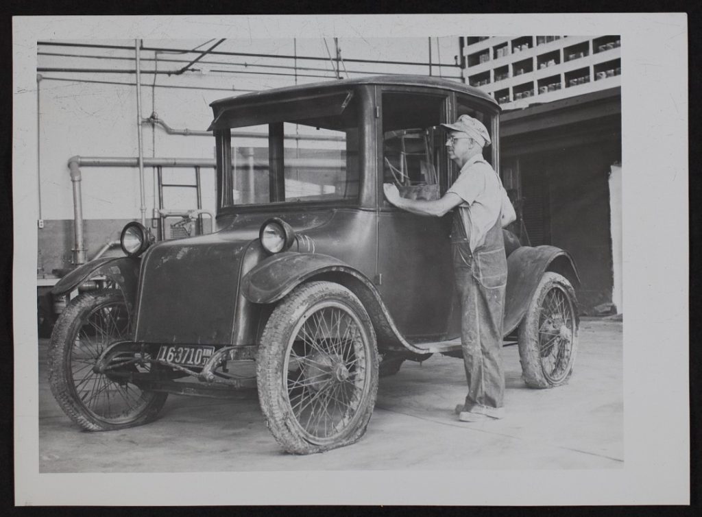 Black-and-white photograph of an older man standing next to a dark colored old-fashioned car.