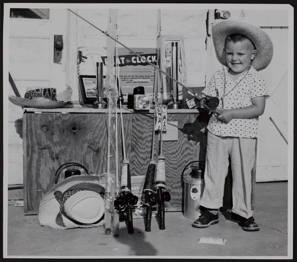 Black-and-white photograph of a young boy standing next to toys displayed on top and in front of a wooden box. The boy is holding a fishing pole.