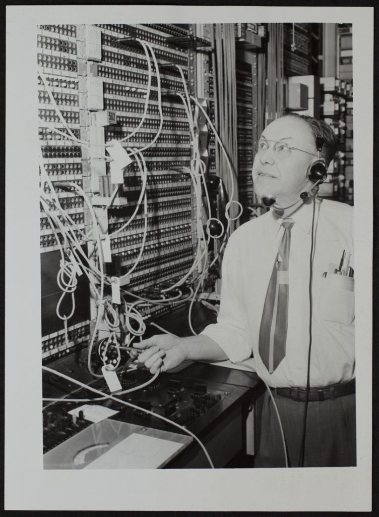 Black-and-white photograph of an older man standing next to a telephone switchboard.