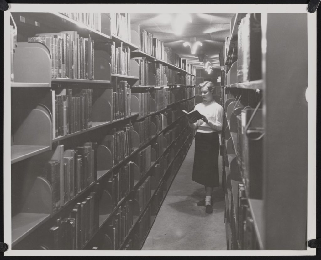 Black-and-white photograph of a women standing and reading between two rows of floor to ceiling bookshelves.