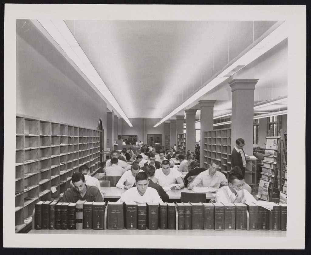 Black-and-white photograph of students sitting and reading at long tables. There are books in the foreground and bookshelves around the parameter of the room.