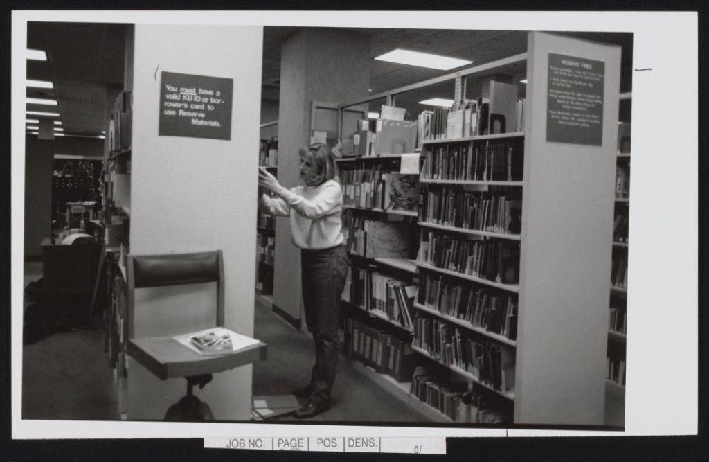 Black-and-white photograph of a woman looking at volumes on a bookshelf.