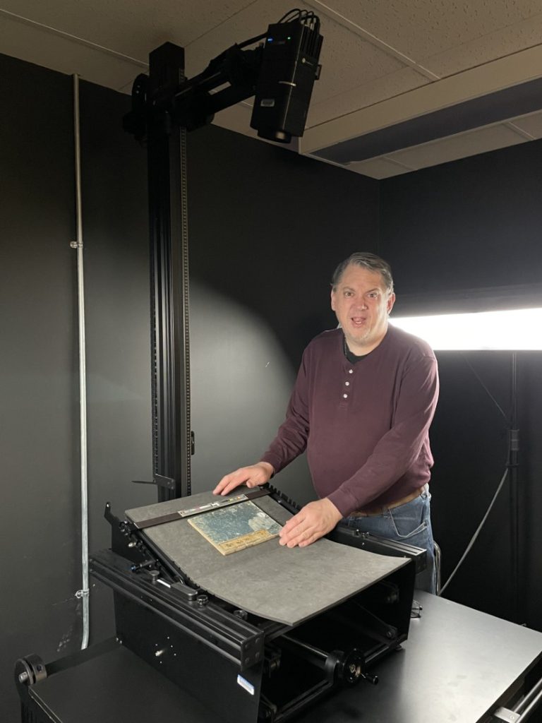 Photograph of a man standing in a darkened room with a book on a raised table and a cylindrical camera looking down from near the ceiling.