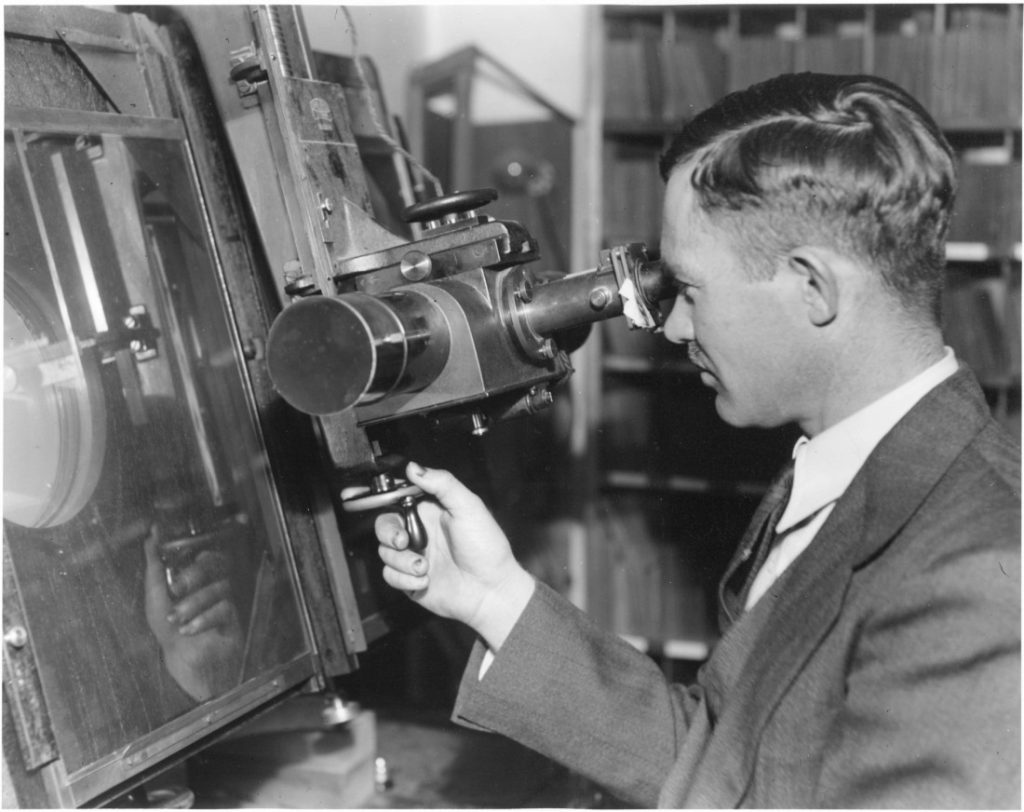 Black-and-white photograph of a man looking through an eyepiece connected to a larger machine.