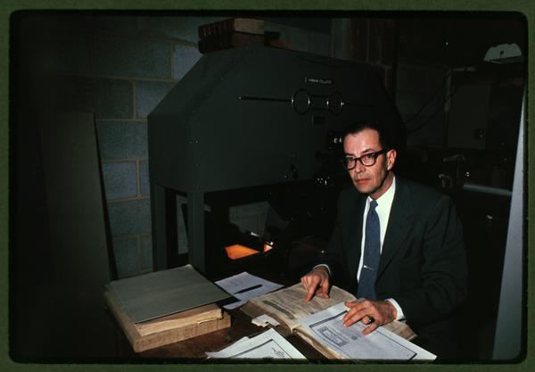 Color photo of a man with glasses sitting at desk covered in books and papers.