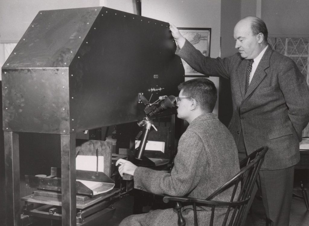 Black-and-white photograph of a man standing in front of a large machine while a second man sits and looks through the eyepiece.