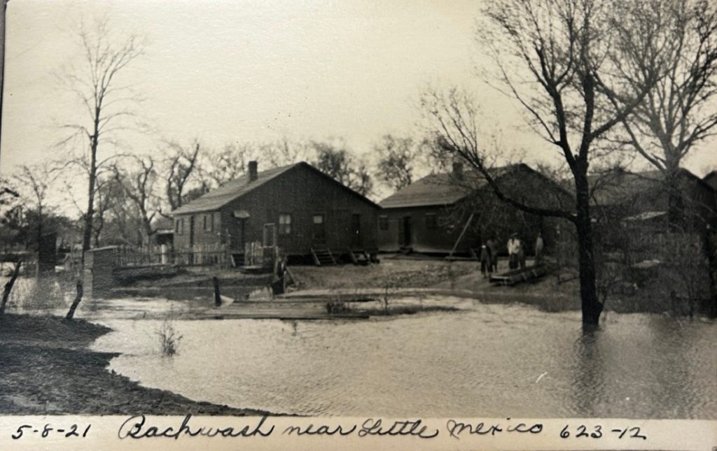 Sepia-toned photograph of a small group of people gathered near standing water with a row of houses in the background.