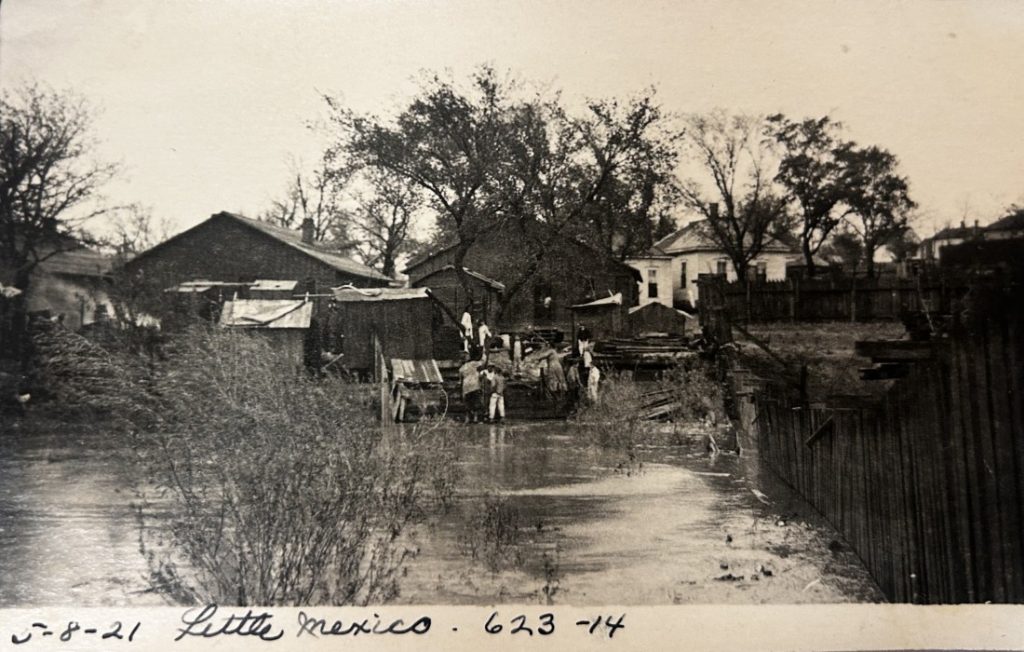 Sepia-toned photograph of people working near floodwaters with houses in the background and large fencing on the right.