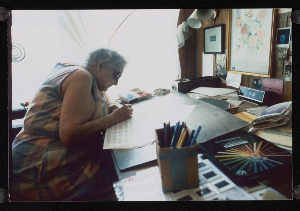 Color photograph of an older woman drawing at a desk with art supplies around her workspace.
