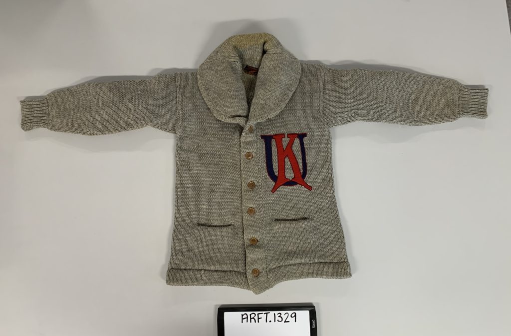 Lela Duncan Cardozo’s KU letter sweater is pictured. The sweater is knitted from gray wool with red capital letter K and blue capital letter U sewn on the left front.