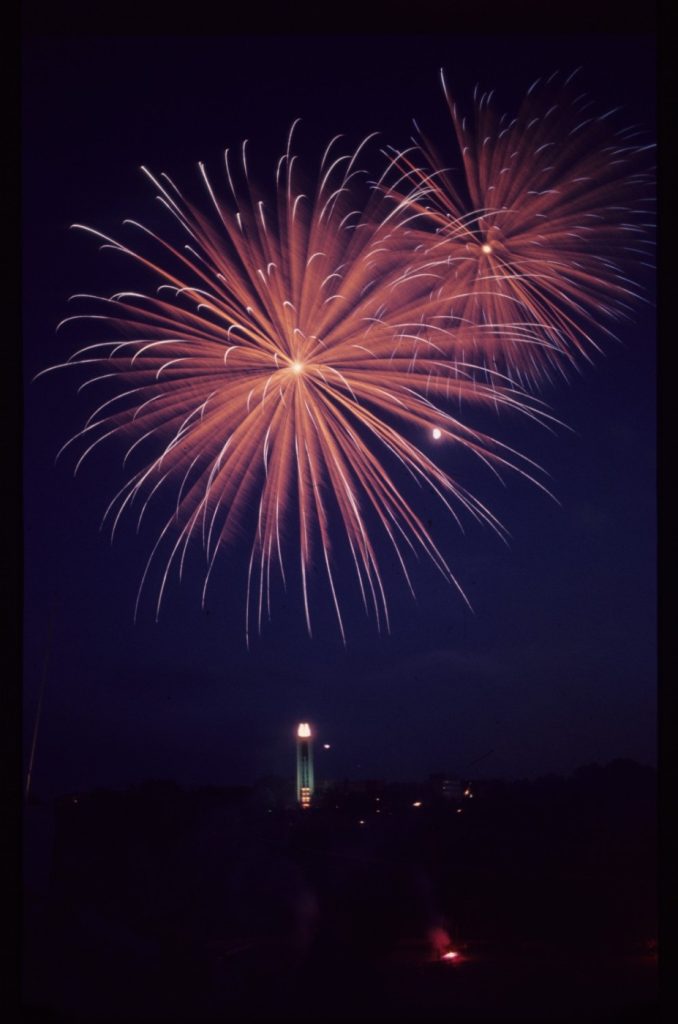 Color photograph of big red fireworks exploding against a dark night sky, with only the light from the Campanile otherwise visible.