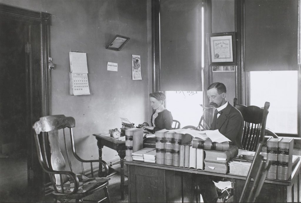 Black-and-white photograph of a man sitting at a desk crowded with books. A woman sits to left, using a typewriter.