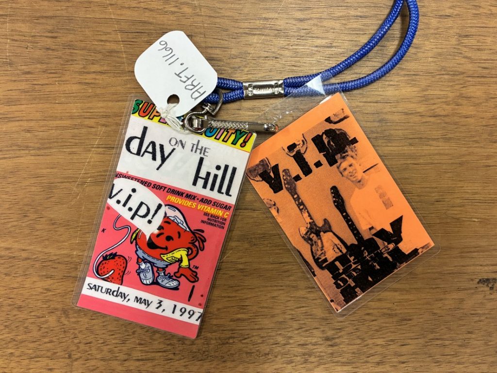 Photo of two VIP badges. One is orange with a background image of a young man and guitars. The other features the Kool-Aid Man.