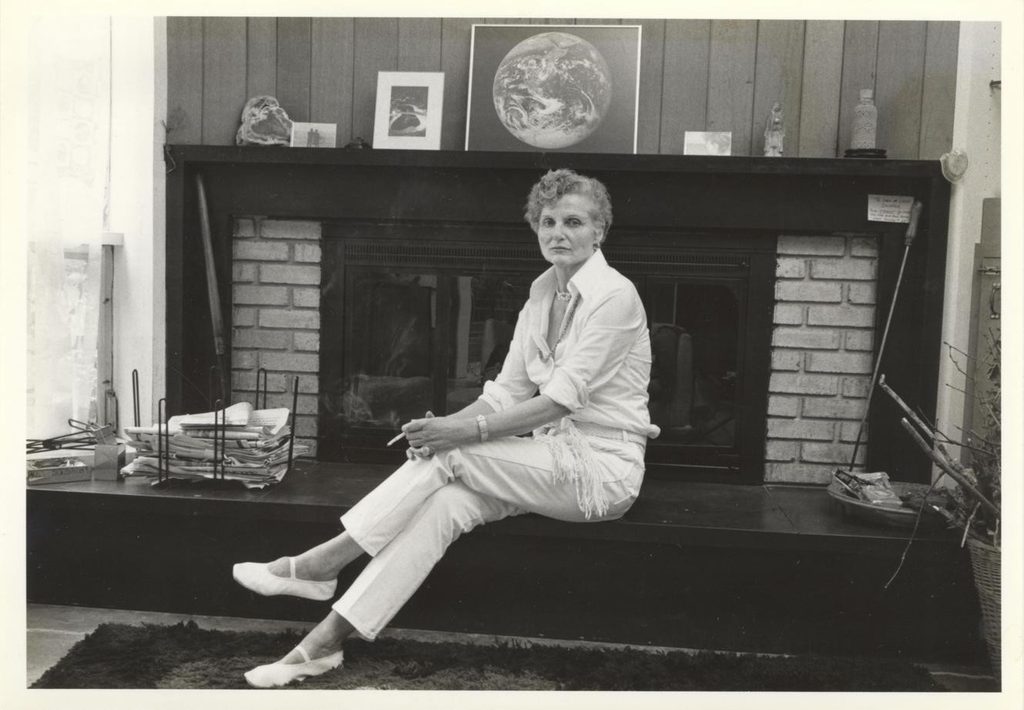 Black and white photograph of author Alice Bradley Sheldon sitting with her legs crossed in front of a fireplace. 